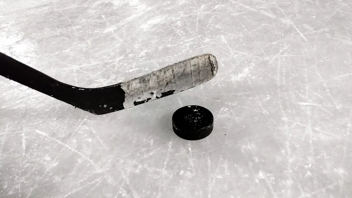 Stick com fita cinzenta encostado a um disco sobre gelo riscado por patins